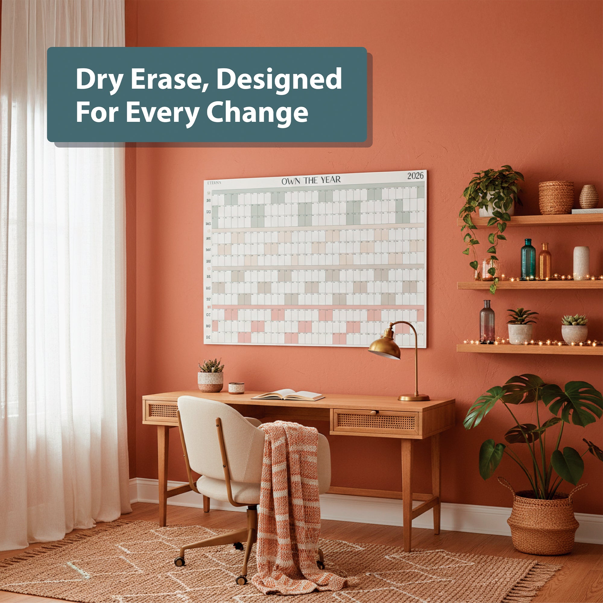 A desk with a chair, lamp, and blanket sits beneath the Eterna Goods "Own the Year 2026 GIANT Dry Erase Wall Calendar" (4ft x 3ft full-year planner) in a tidy home office with shelves and plants.