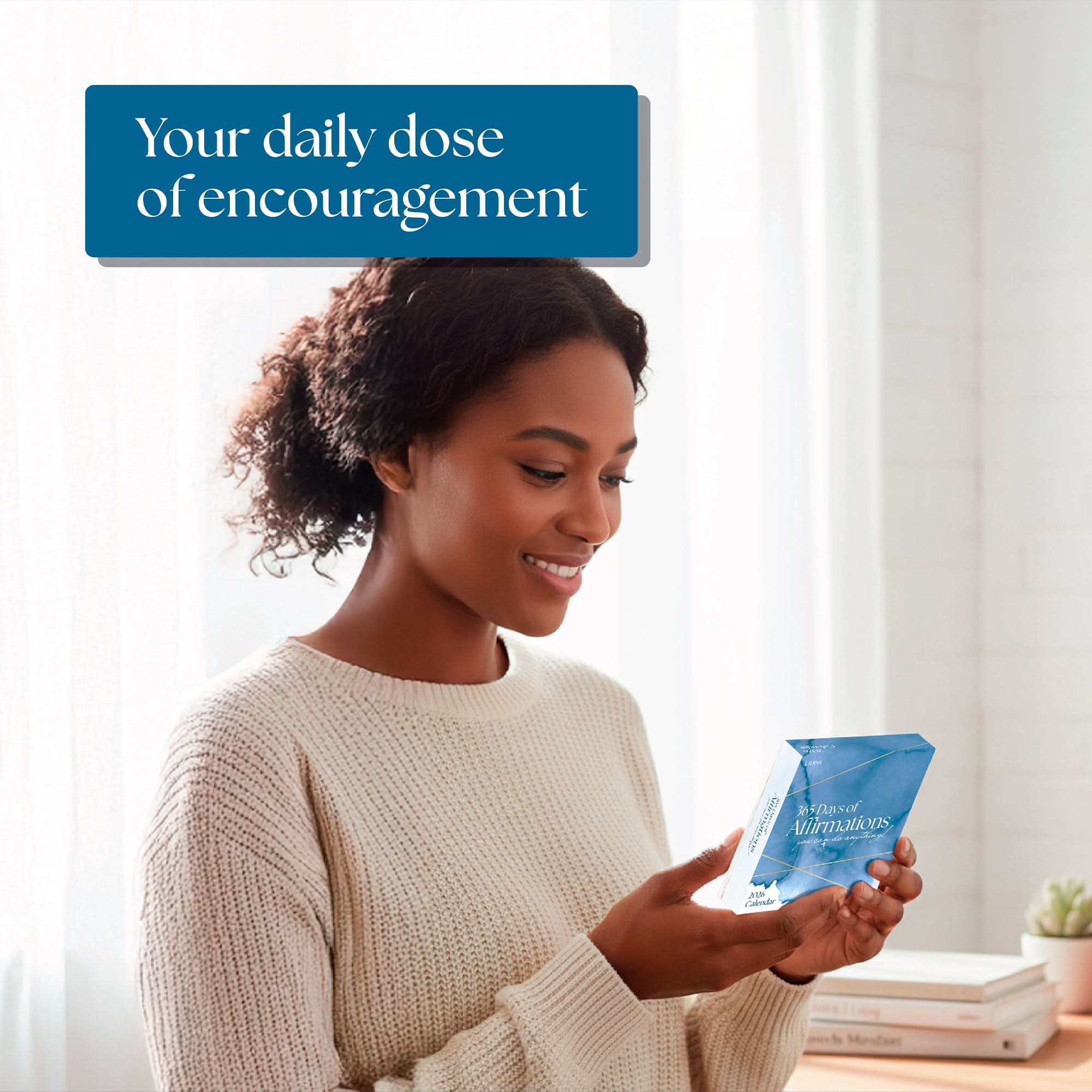 A woman in a cream sweater smiles while reading the blue “2026 Daily Affirmations Calendar” by Eterna Goods in a bright, softly lit room. Text above reads, “Your daily dose of encouragement.”.