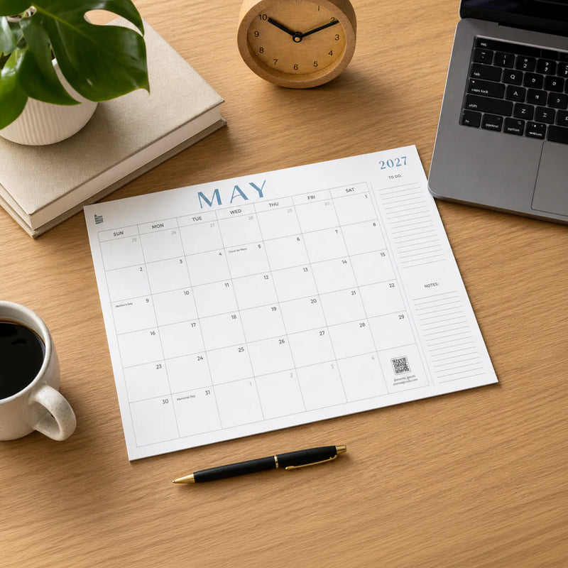 A blank August 2026 wall calendar hangs on a gray concrete wall above a white table and wooden chairs, with a small green plant in the foreground.