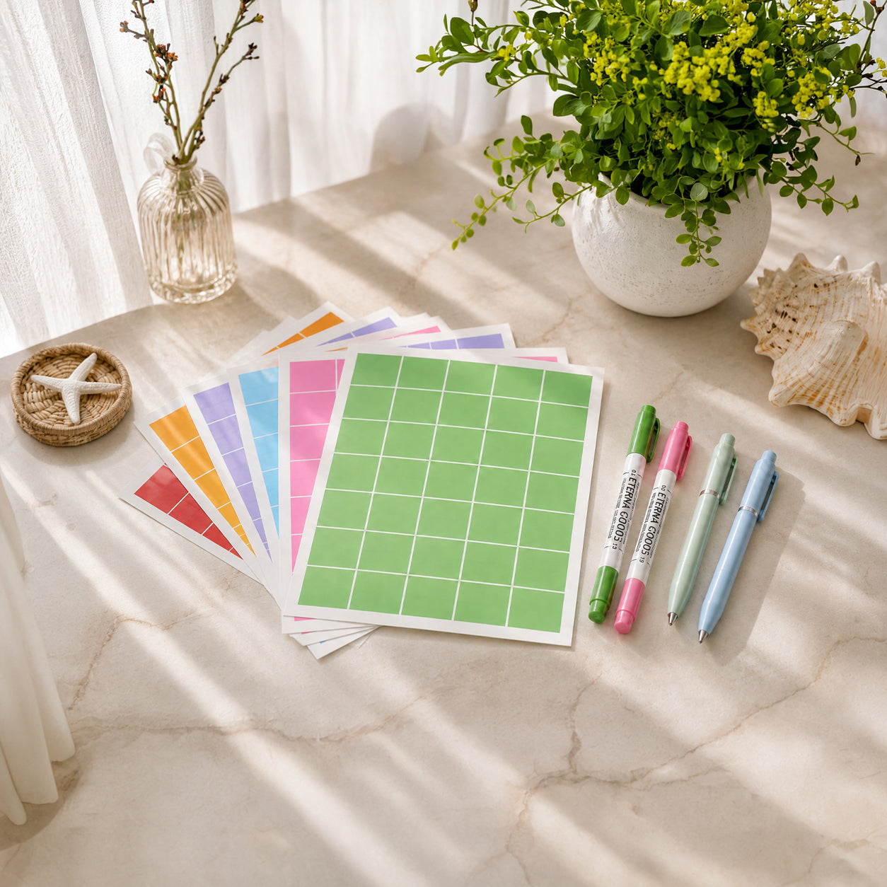 Several sheets of colorful grid sticker paper and four pastel pens are arranged on a marble surface next to a potted plant, a small vase, and seashell decor.