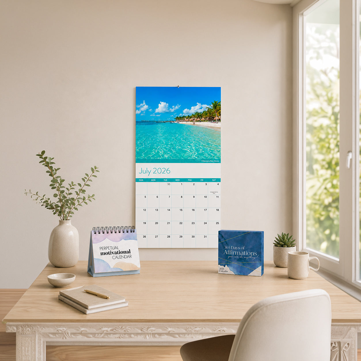 A neatly arranged desk with a potted plant, stationery, two calendars, and a cup, with a wall calendar showing July 2026 and a beach scene hanging above.