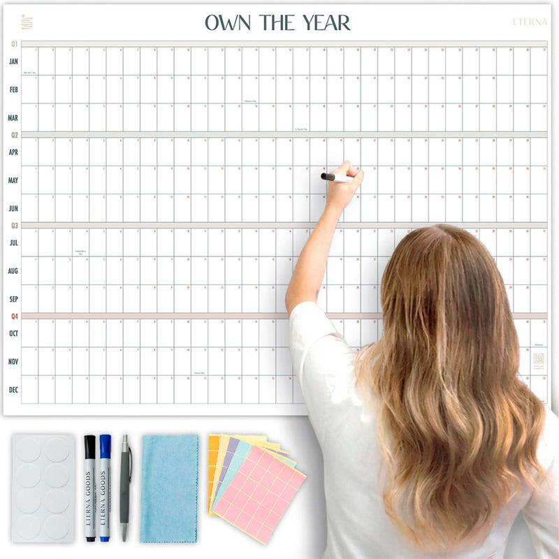 A person writes on the Eterna Goods Own the Year Undated GIANT Dry Erase Wall Calendar (4ft x 3ft); markers, an eraser, and stickers are displayed below this full year planner.