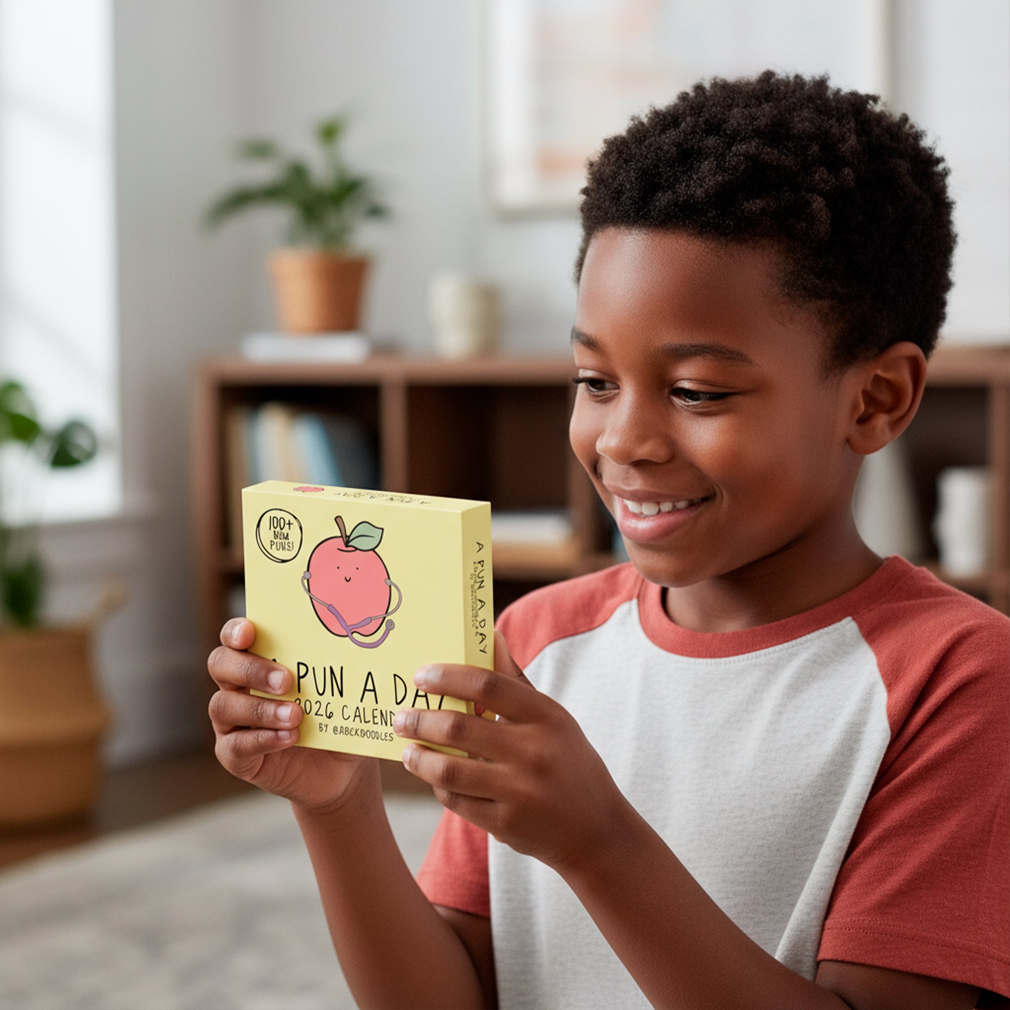A smiling boy holds and looks at the 2026 A Pun a Day Calendar by rockdoodles, featuring a peach illustration on the front, in a cozy living room setting.