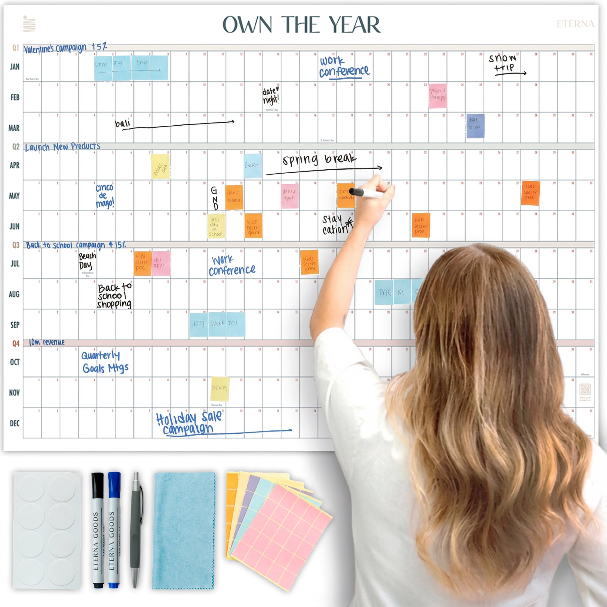 A woman writes on the Eterna Goods Own the Year UNDATED GIANT Dry Erase Wall Calendar (4ft x 3ft) using a marker; markers, eraser, magnets, and sticky notes are at the bottom left.