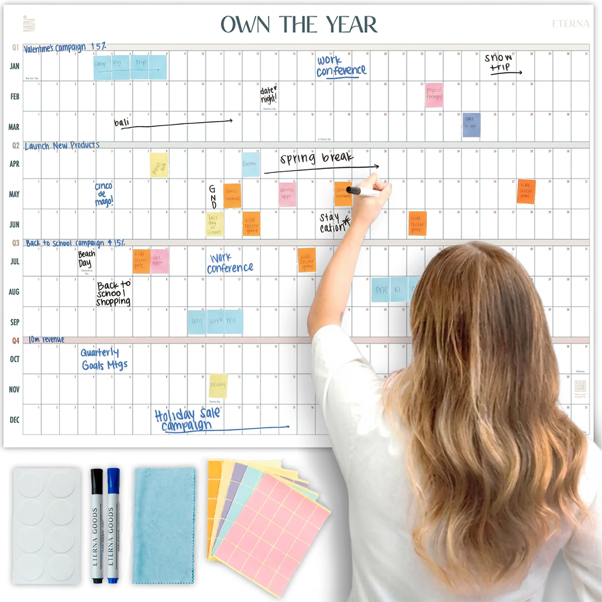 A woman writes on the Eterna Goods Own the Year UNDATED GIANT Dry Erase Wall Calendar (4ft x 3ft) with colorful markers, stickers, and wipes shown below; events and notes in various colors fill the full year planner.