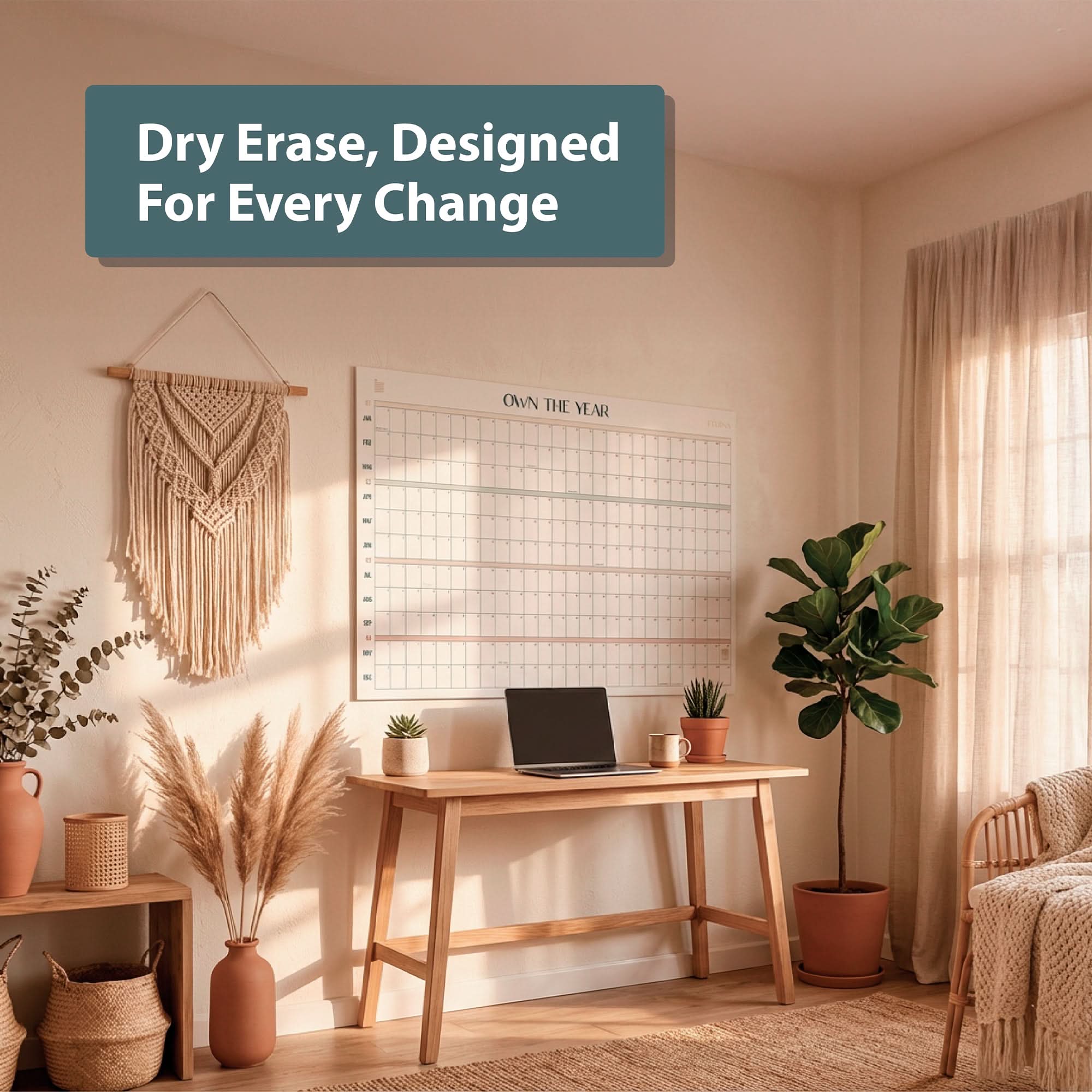A minimalist home office features a wooden desk, laptop, potted plants, and the Eterna Goods Own the Year Undated GIANT Dry Erase Wall Calendar (4ft x 3ft) on the wall. Sunlight pours in through sheer curtains.