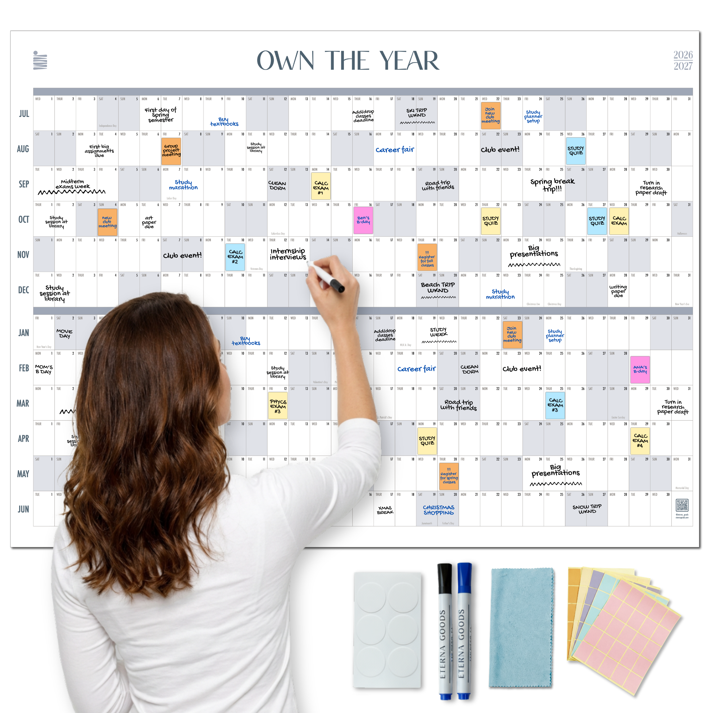 A woman stays organized with the Eterna Goods Midyear/Academic Own the Year 2026-2027 GIANT Dry Erase Wall Calendar (4ft x 3ft), full of color-coded events, using included markers, eraser, cleaning cloth, and sticky notes.