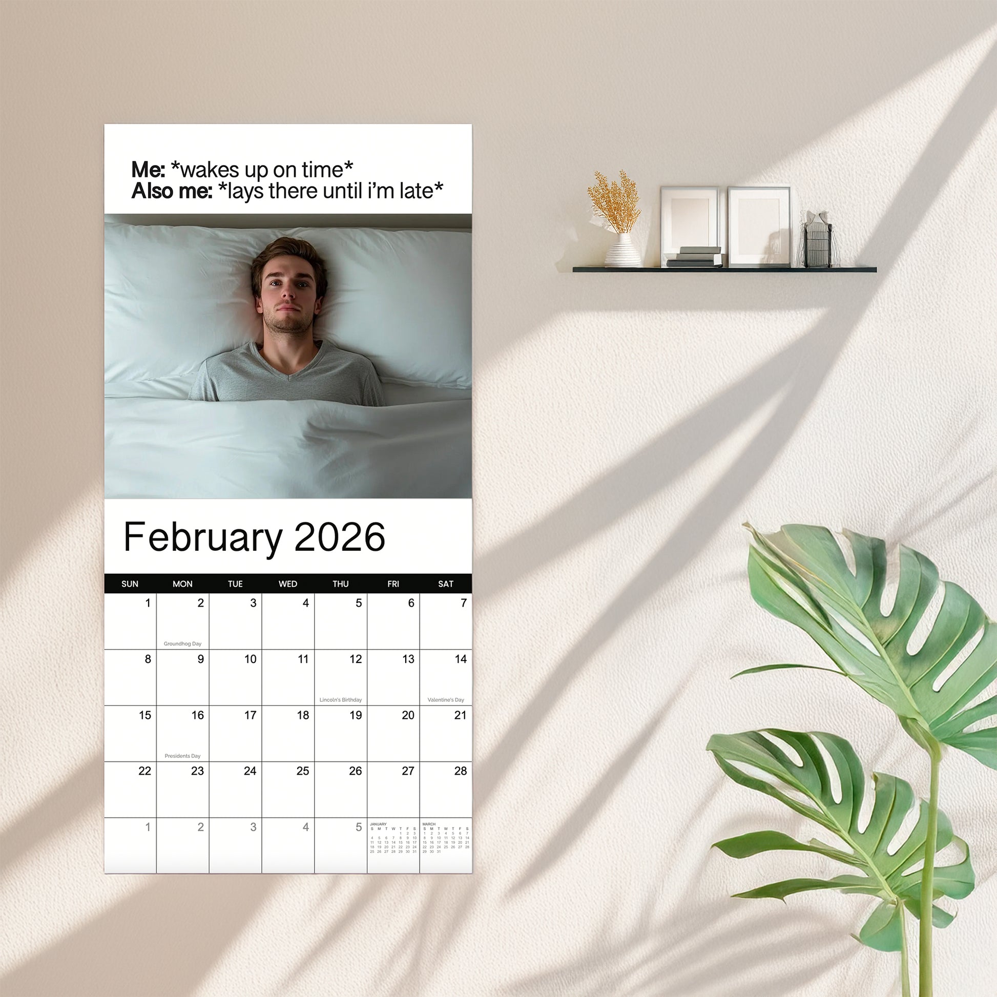A 2026 Meme Wall Calendar by Eterna Goods hangs on the wall, showing a photo of a man in bed with humor about waking up on time but staying in bed. Nearby, there’s a shelf and a plant.