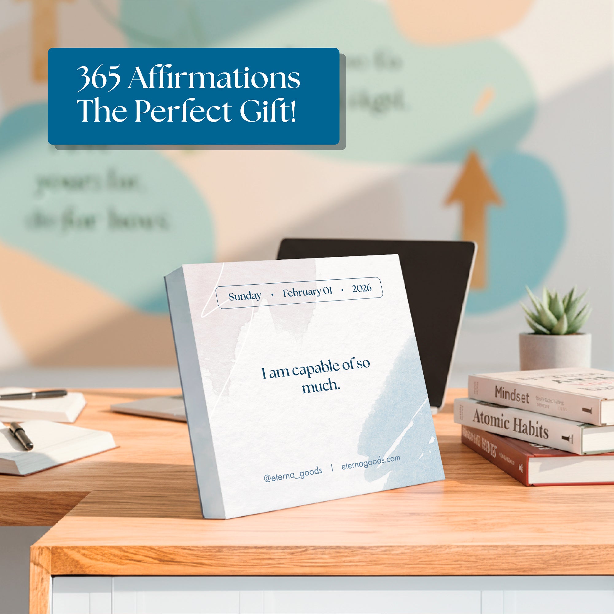 A desk featuring the Eterna Goods 2026 Daily Affirmations Calendar displaying "I am capable of so much," surrounded by books, a plant, and office supplies; a blurred motivational poster appears in the background.
