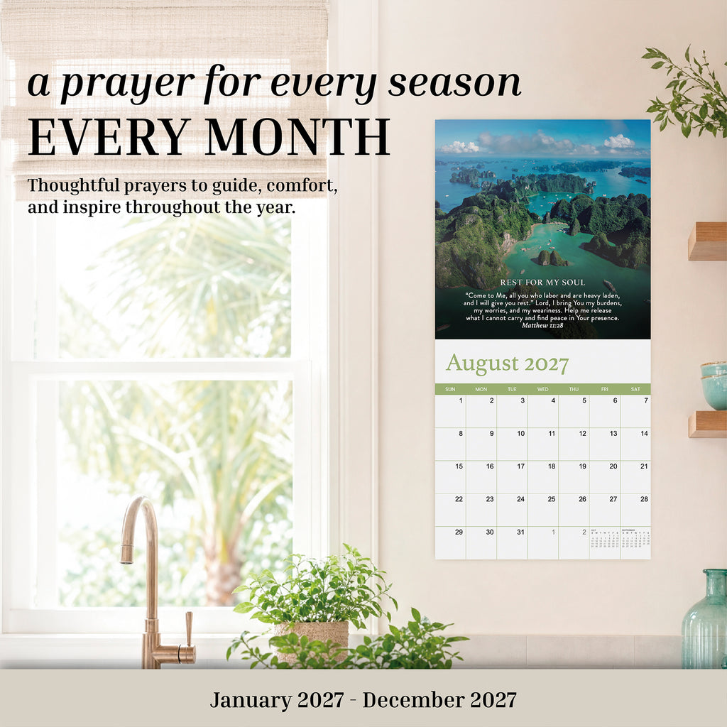 The 2027 Prayer Wall Calendar by Hannah Faith hangs above a wooden table adorned with plants, books, and a lamp. Open to August 2027, this calendar showcases a beautiful landscape photo and an uplifting prayer, making it an inspiring Bible verse calendar—ideal as a thoughtful faith-based gift.