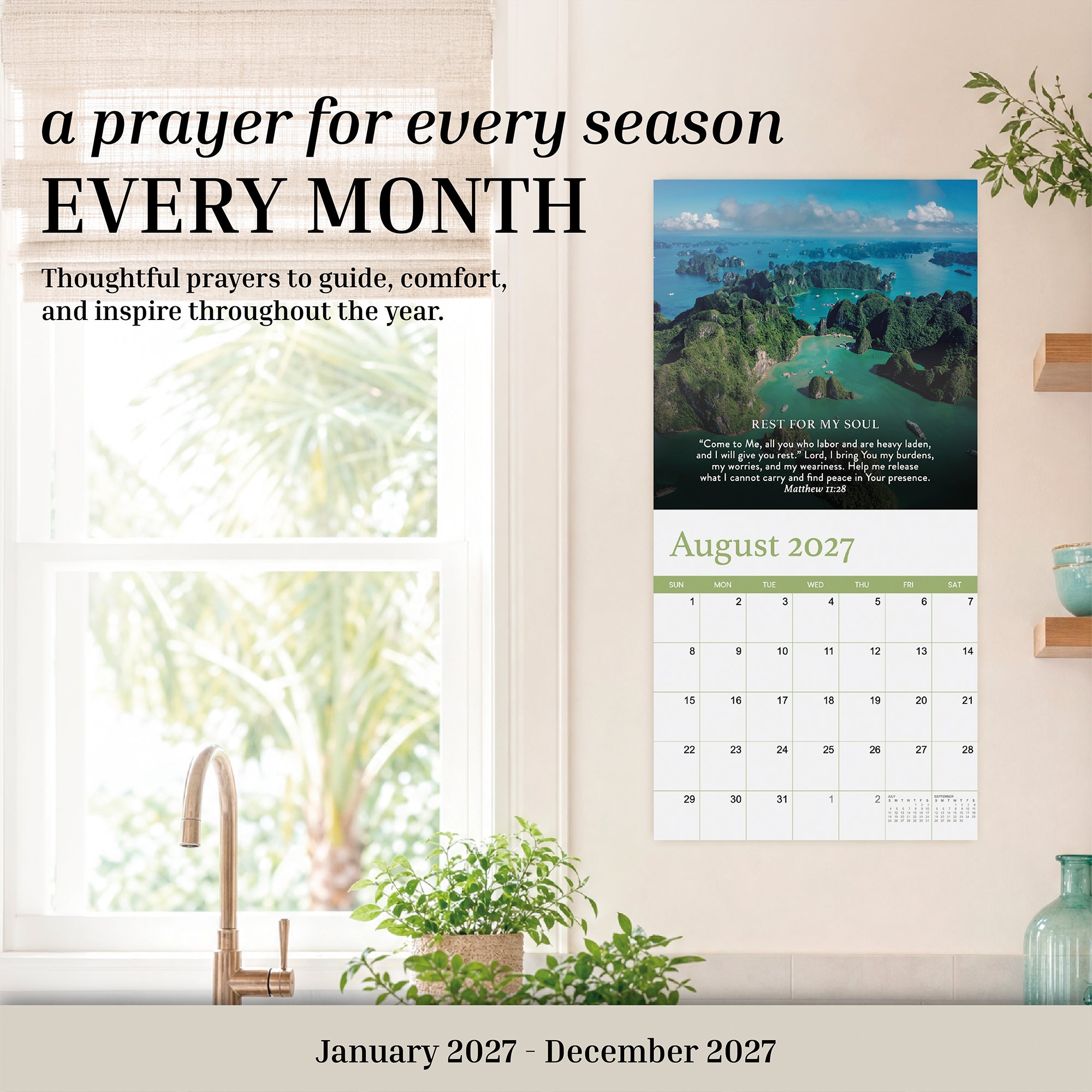 The 2027 Prayer Wall Calendar by Hannah Faith hangs above a wooden table adorned with plants, books, and a lamp. Open to August 2027, this calendar showcases a beautiful landscape photo and an uplifting prayer, making it an inspiring Bible verse calendar—ideal as a thoughtful faith-based gift.