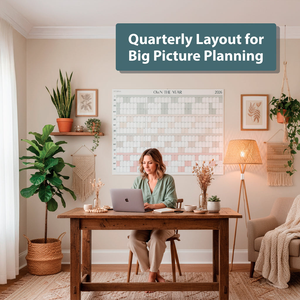 A woman works on a laptop at a wooden desk in a decorated home office with the Eterna Goods "Own the Year 2026" GIANT Dry Erase Wall Calendar (4ft x 3ft) displayed for big picture planning.