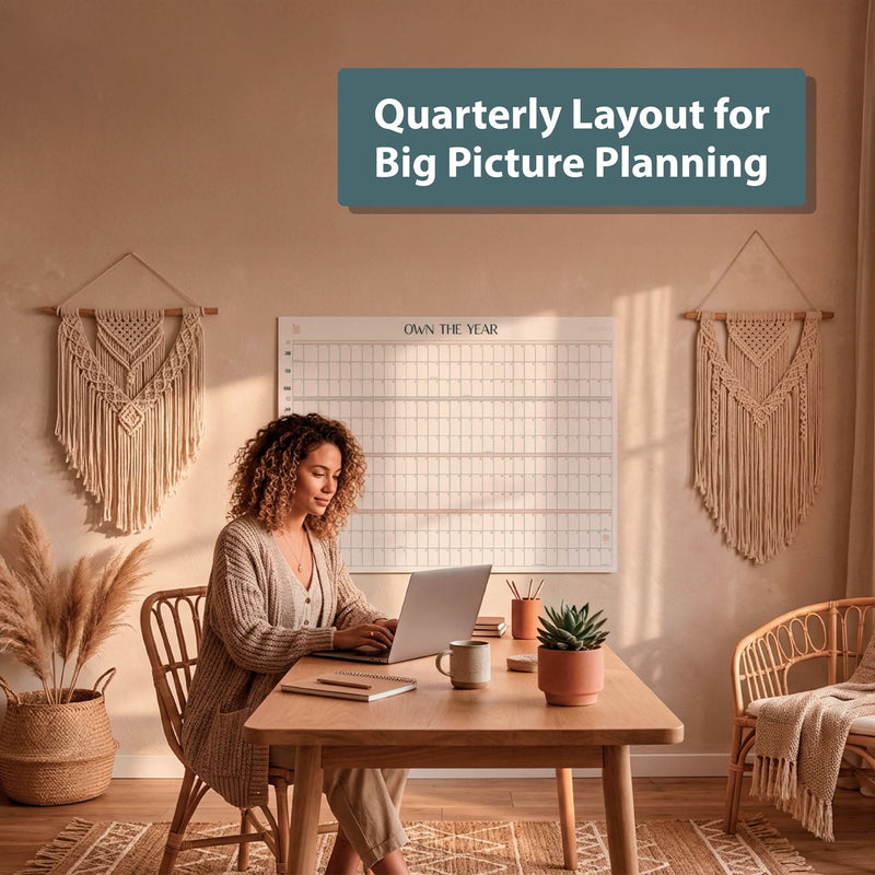 A woman works on a laptop at a wooden desk in a decorated room with an Eterna Goods Own the Year Undated GIANT Dry Erase Wall Calendar (4ft x 3ft) for big picture planning.