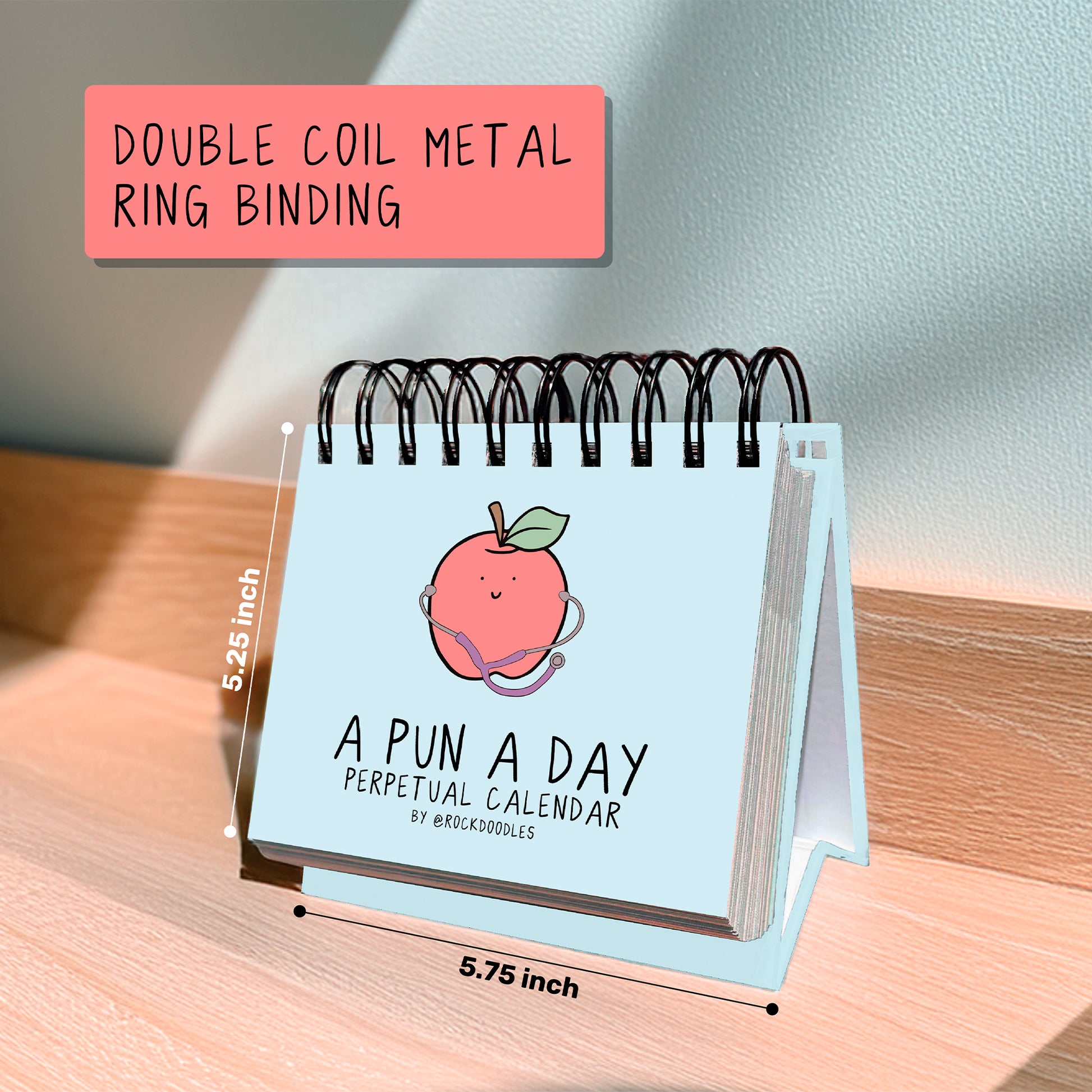 The "A Pun A Day Perpetual Calendar" by rockdoodles, a small double coil metal ring-bound desk calendar (5.25 x 5.75 in), is displayed on a wooden surface.