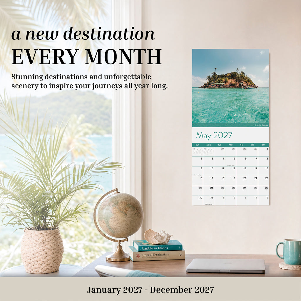 A home office with a desk, chair, and tropical decor features the Eterna Goods 2027 Travel Inspiration Wall Calendar open to May, displaying an island photograph and inspiring text introducing a new travel destination each month.