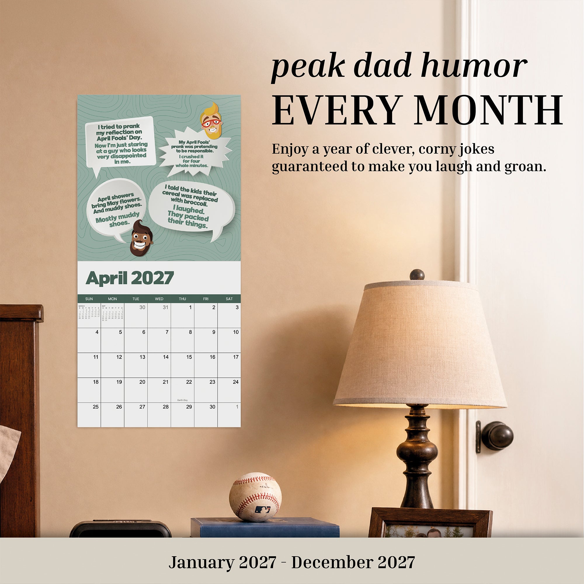 A 2027 Dad Joke Wall Calendar by That Is So Dad, titled "Peak Dad Humor," hangs above a lamp and table with a baseball, featuring funny dad jokes and cartoon graphics for each month.