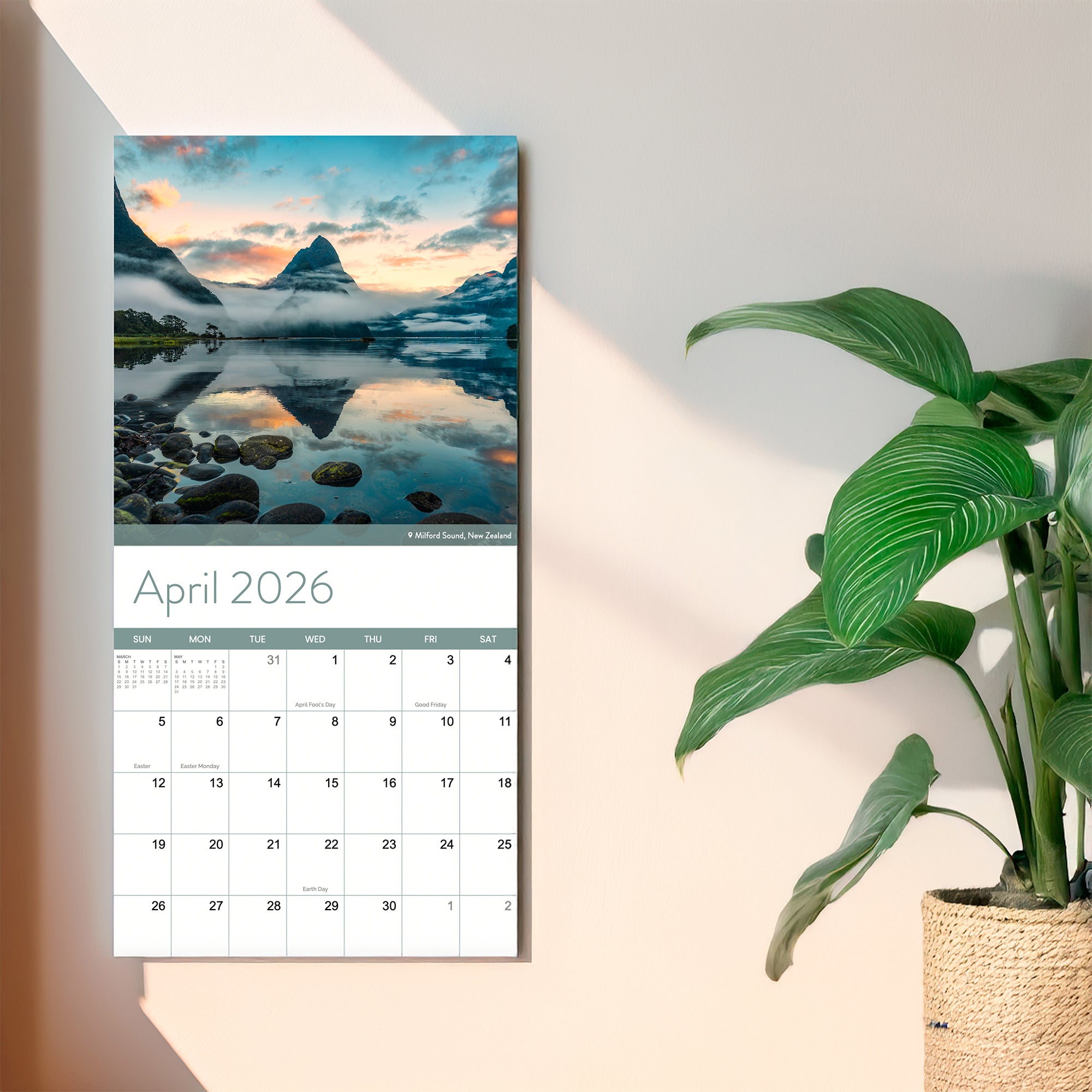 An Eterna Goods 2026 Travel Inspiration Wall Calendar, open to April and showcasing breathtaking Milford Sound, New Zealand, coastal photography, hangs above a lush potted plant in a woven basket.