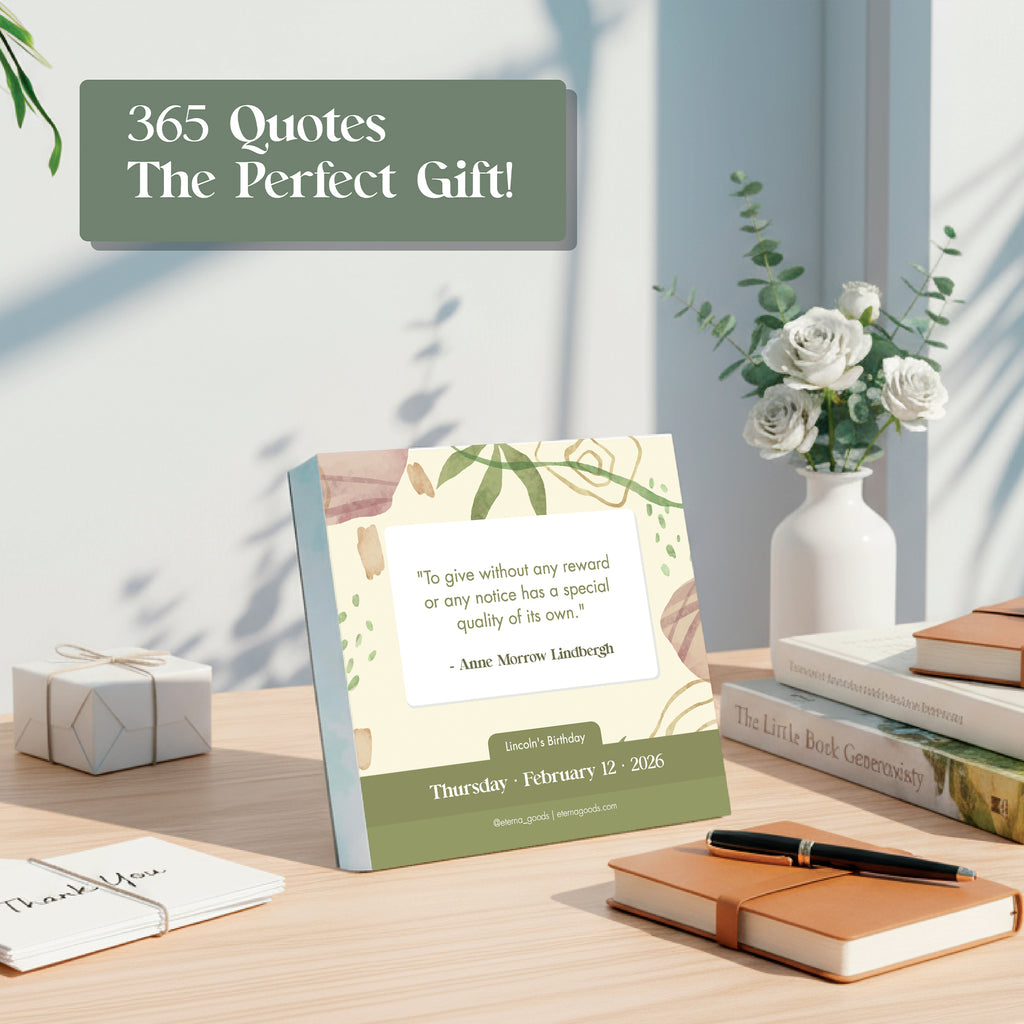 A desk featuring the Eterna Goods 2026 Motivational Calendar displaying a quote, surrounded by white flowers in a vase, books, a pen, and gift boxes.