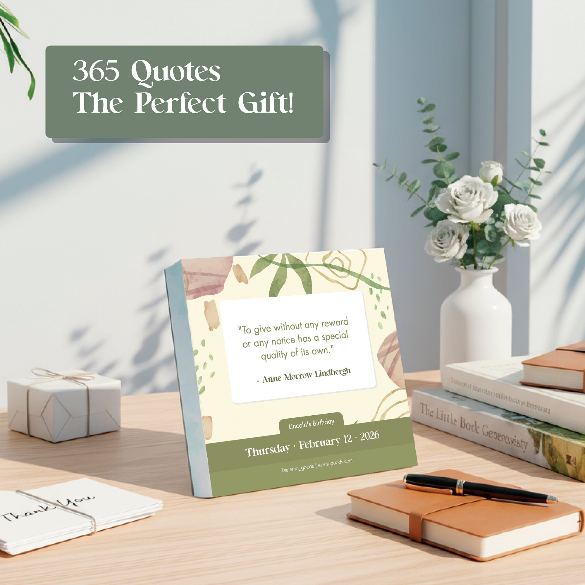 A desk featuring the Eterna Goods 2026 Motivational Calendar displaying a quote, surrounded by white flowers in a vase, books, a pen, and gift boxes.