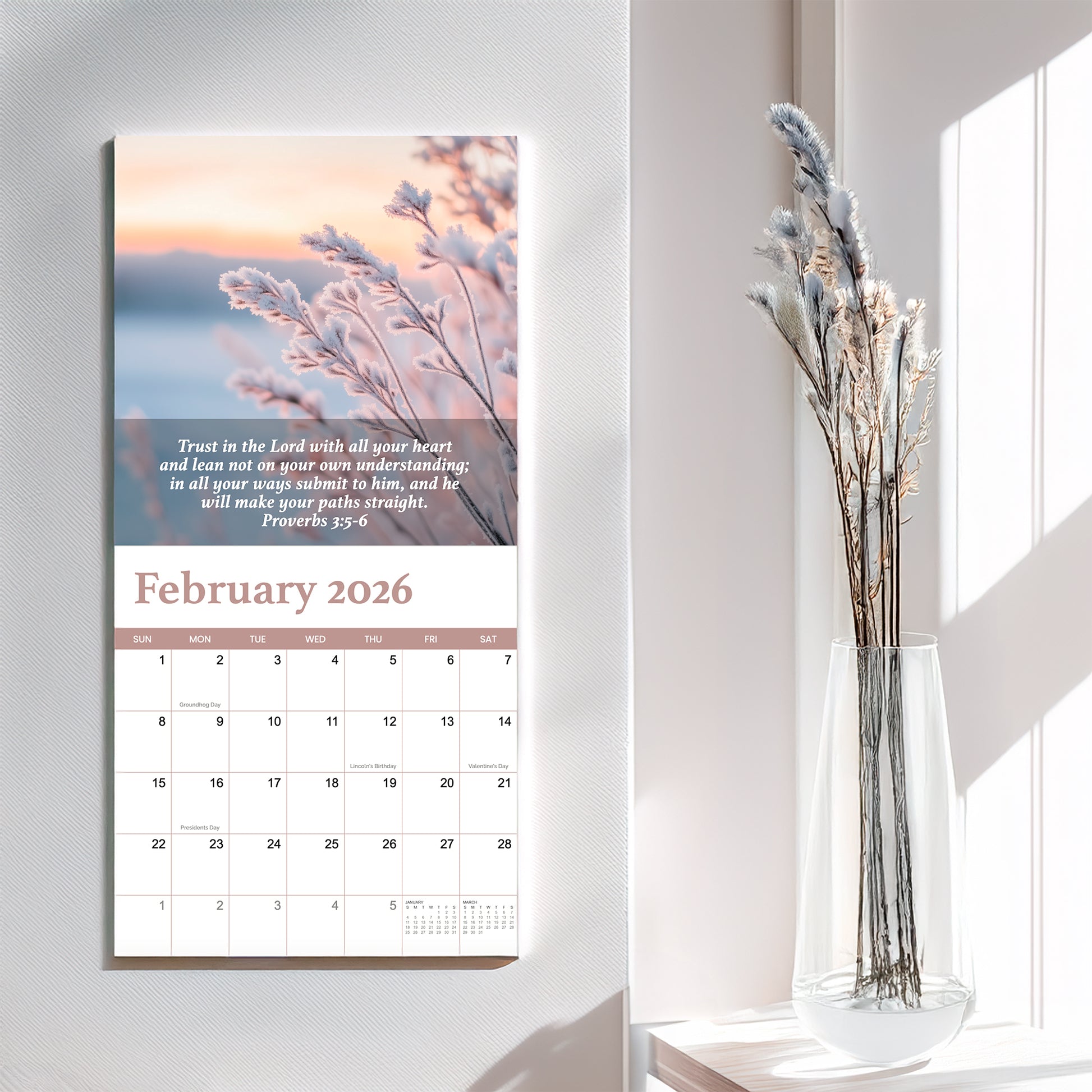 A 2026 Bible Verse Wall Calendar by Hannah Faith, open to February and displaying a photo of frosted plants in gentle light and an inspiring verse, hangs beside a vase of dried flowers.