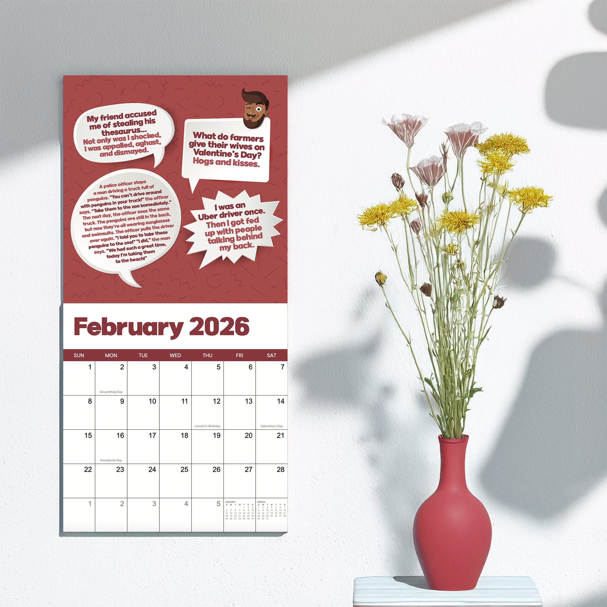 A 2026 Dad Joke Wall Calendar by That Is So Dad hangs on a white wall next to a red vase of yellow and pink flowers. The calendar displays jokes and speech bubbles at the top.