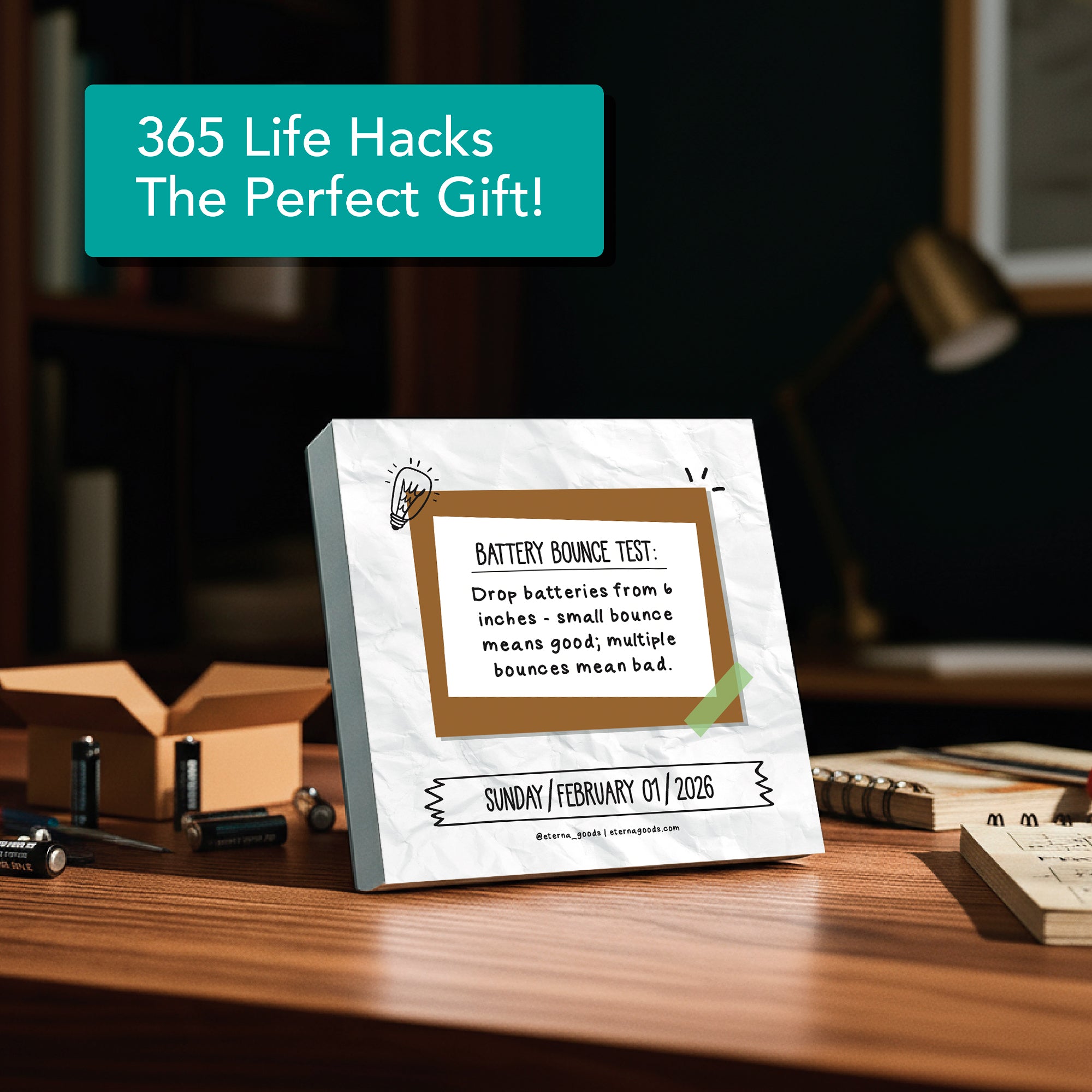 The Eterna Goods 2026 Daily Life Hacks Calendar sits on a desk with batteries and books, displaying a tip about testing batteries by bouncing them—a practical gift for any workspace.