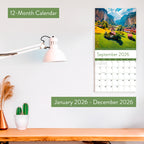 The Eterna Goods 2026 Travel Inspiration Wall Calendar, with stunning coastal photography and January–December 2026 coverage, hangs above a desk decorated with a lamp, small plant, and cactus opened to September.