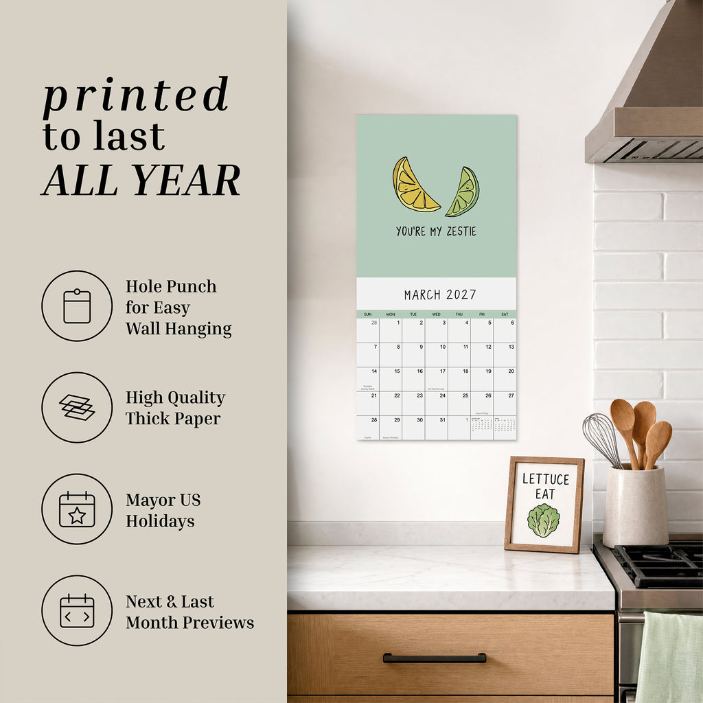 A kitchen showcasing the rockdoodles 2027 Pun Wall Calendar for March—decorated with playful lemon artwork—hangs beside a framed print of lettuce, with utensils neatly organized in a jar. Promotional text highlights the calendar’s unique features, emphasizing that this funny and cheerful gift from rockdoodles is perfect for pun enthusiasts looking to add humor and brightness to their space.