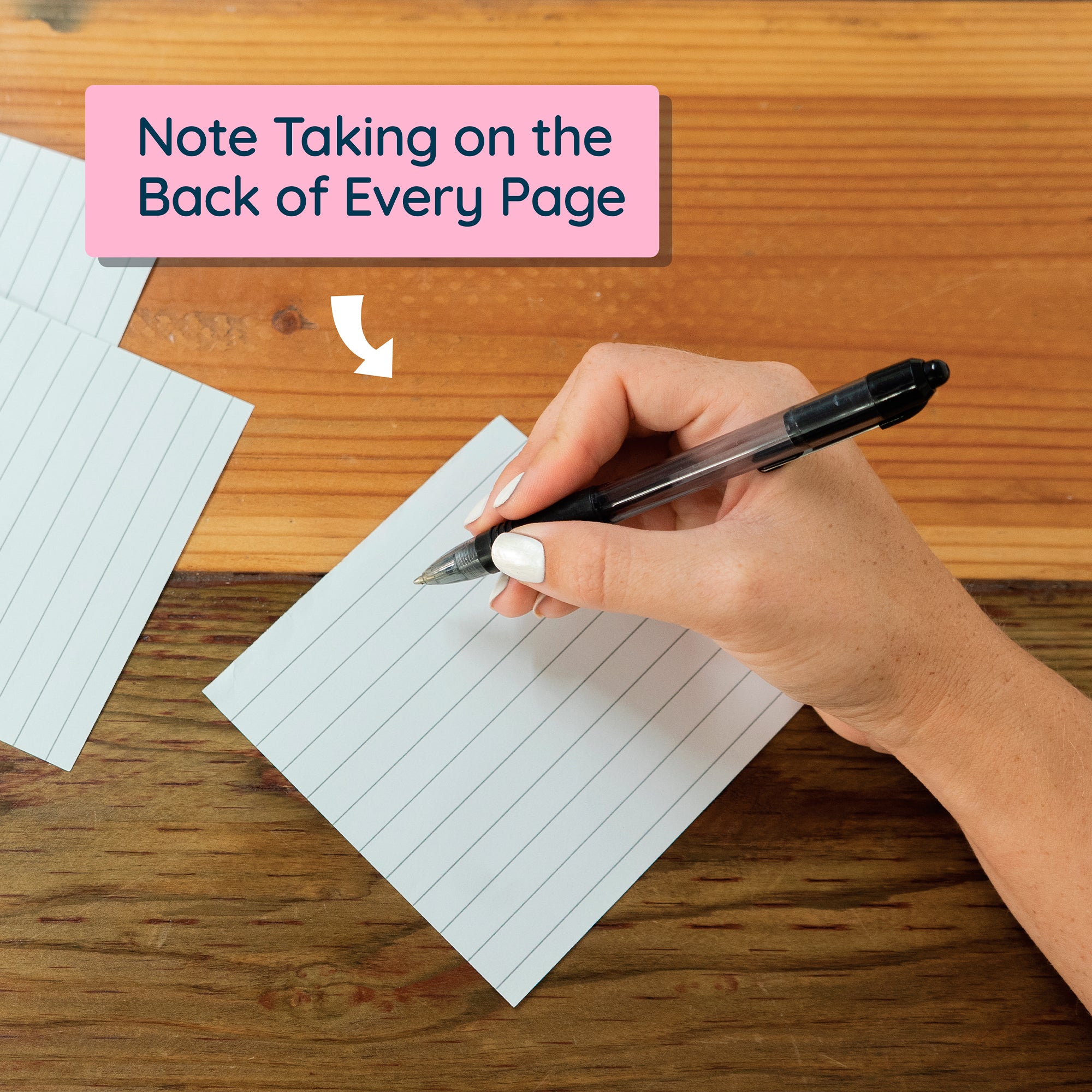A hand holds a black pen, writing on the back of a lined note card on a wooden table. “Note Taking on the Back of Every Page”—ideal for your Eterna Goods 2026 Word Of The Day Calendar or vocabulary builder routine.
