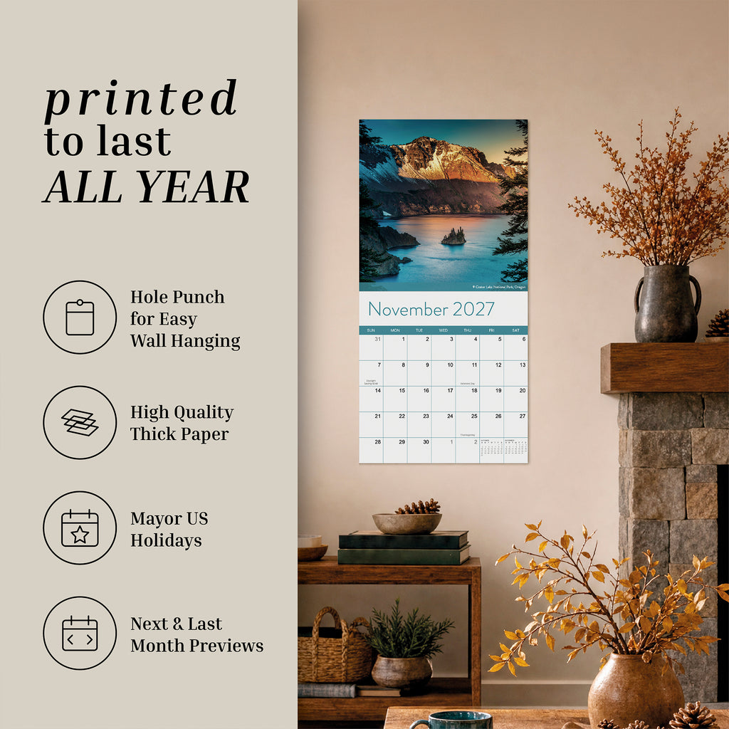 An Eterna Goods 2027 National Parks Wall Calendar hangs in the living room above a shelf displaying books, a plant, and autumn-inspired decor; ideal for outdoor enthusiasts, this calendar showcases breathtaking National Park photography with a mountain lake scene and product highlights.