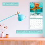 The 2026 Dogs Wall Calendar by Eterna Goods hangs above a desk, open to June, featuring coastal photography and a playful image of a dog swimming underwater.