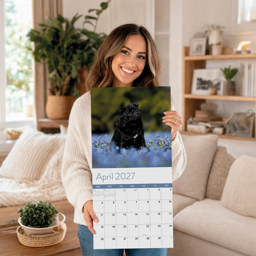 A woman holds the Eterna Goods 2027 Dogs Wall Calendar opened to April, showcasing a breathtaking photo of a black dog sitting in a field of blue flowers—an ideal calendar for any dog lover.