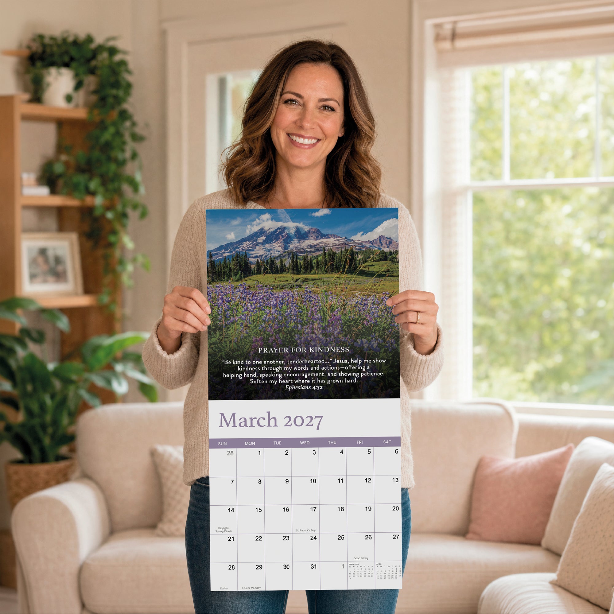 A woman stands smiling in her living room, holding up the Hannah Faith 2027 Prayer Wall Calendar, opened to March and displaying a breathtaking mountain and wildflower landscape with a prayer for kindness—an inspiring faith-based gift designed to bring daily beauty and encouragement.