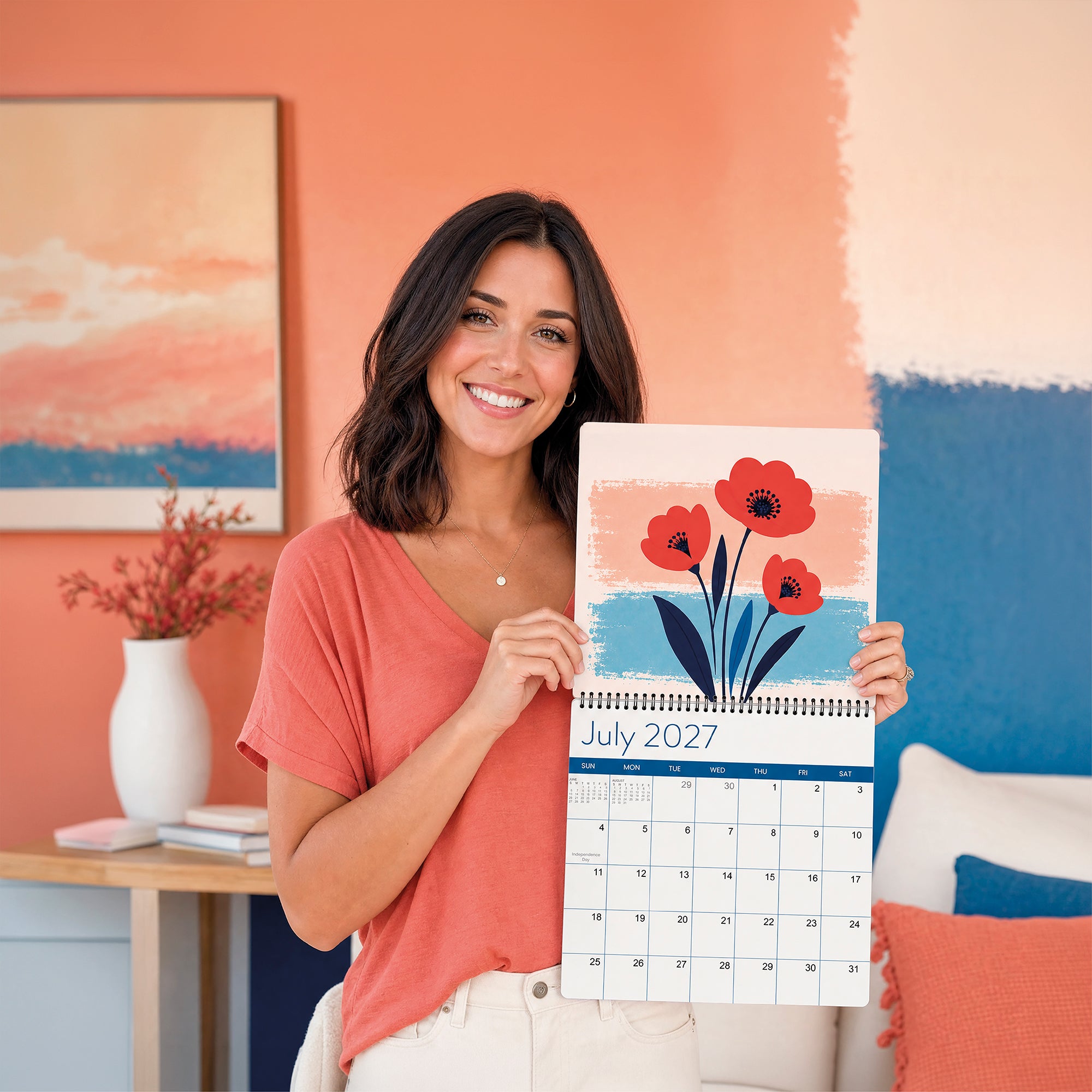 A woman holds open the Eterna Goods 2027 Floral Wall Calendar to July, displaying a vibrant floral illustration; behind her are orange and blue wall décor and a painting.