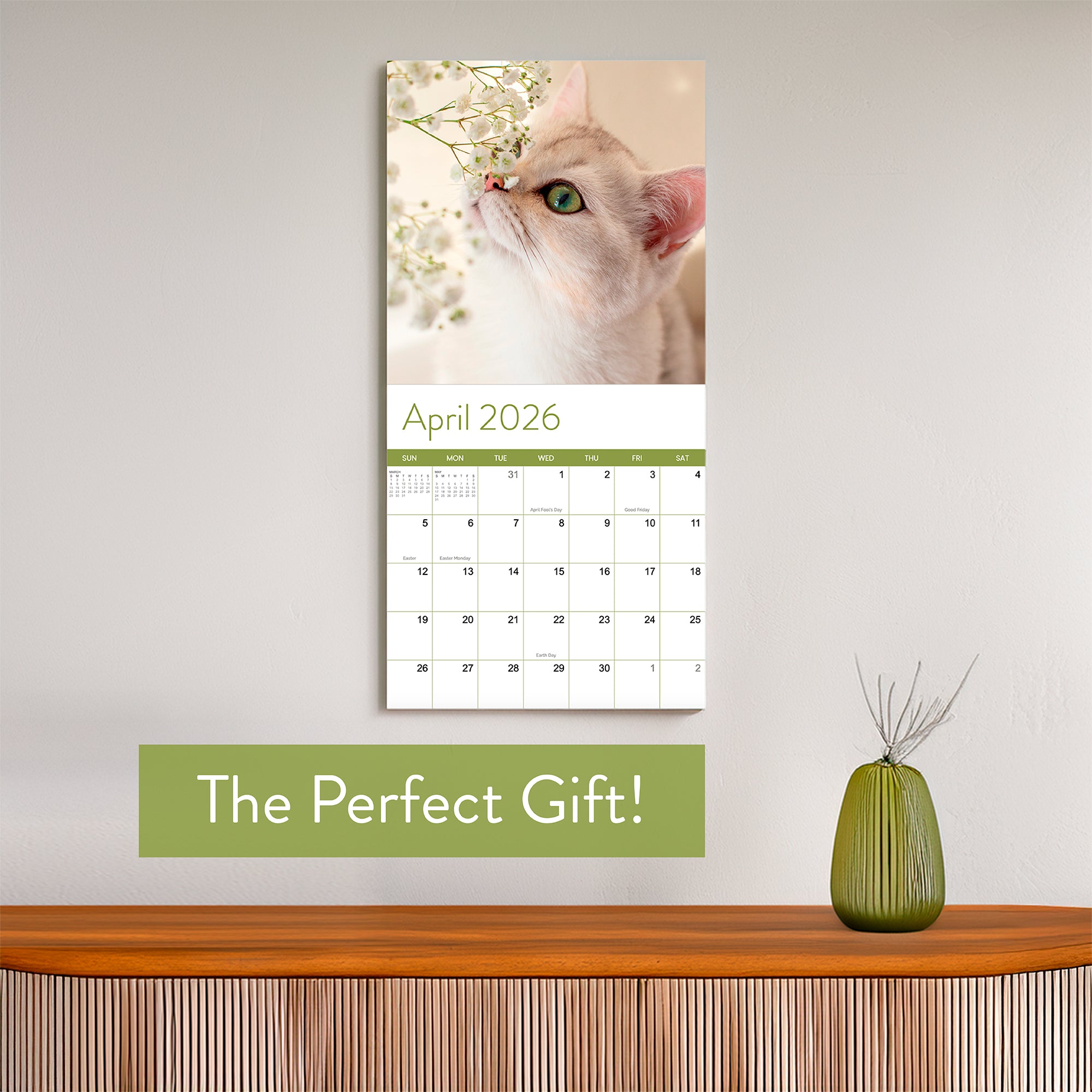 The 2026 Cats Wall Calendar by Eterna Goods features an April page with a cat sniffing white flowers and "The Perfect Gift!" on a green banner.