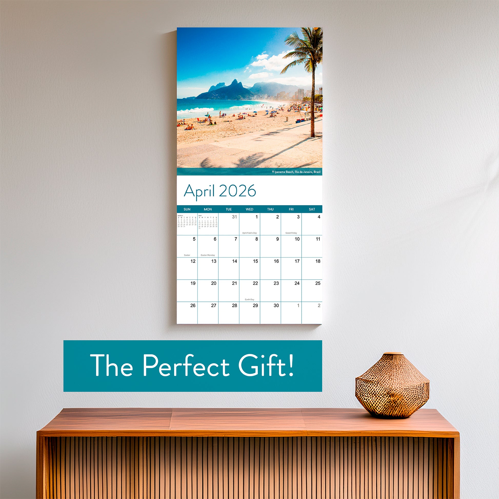 The Eterna Goods 2026 Beaches Wall Calendar, open to April and displaying a beach scene, hangs above a wooden console table with a wicker vase. Text reads: "The Perfect Gift!.