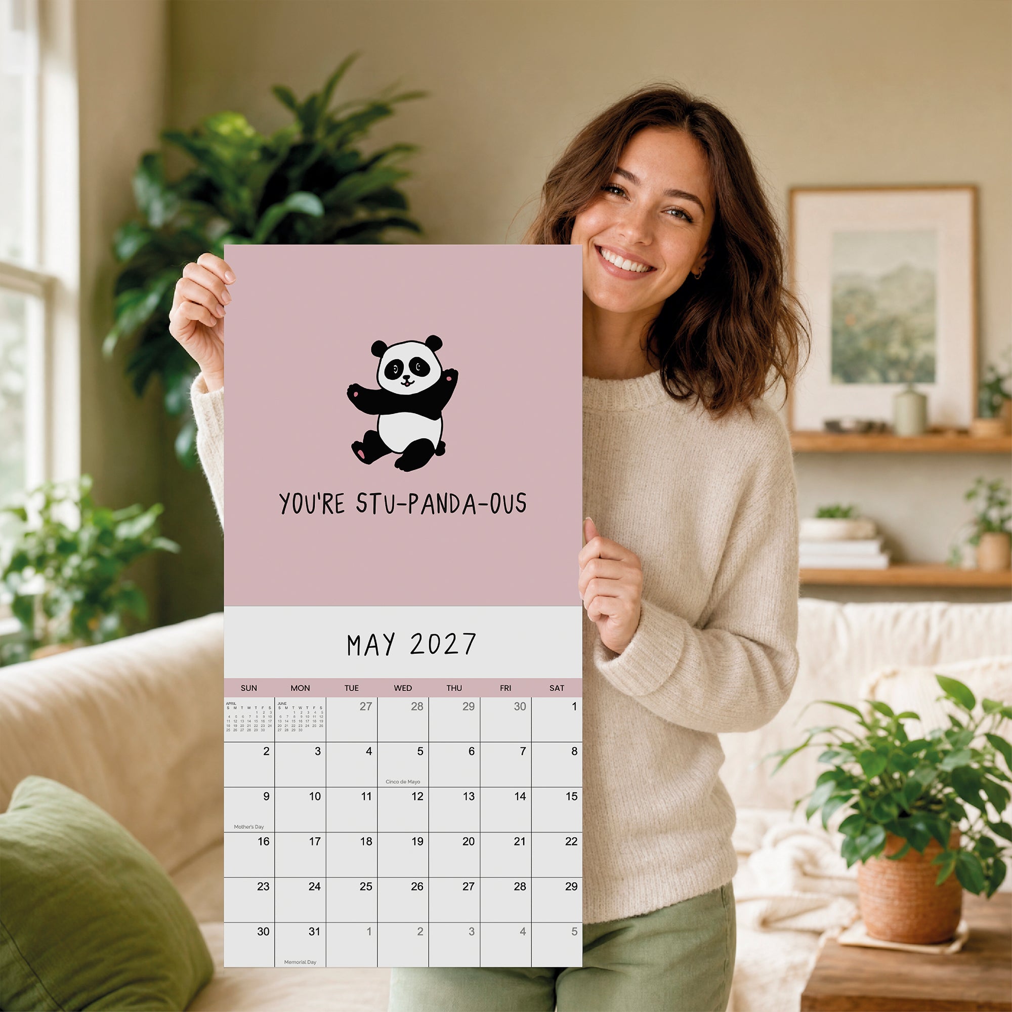 A woman holds a large rockdoodles 2027 Pun Wall Calendar in a cozy living room filled with plants and framed art, displaying a charming cartoon panda and the phrase “You’re stu-panda-ous”—an ideal funny calendar and gift for pun enthusiasts.