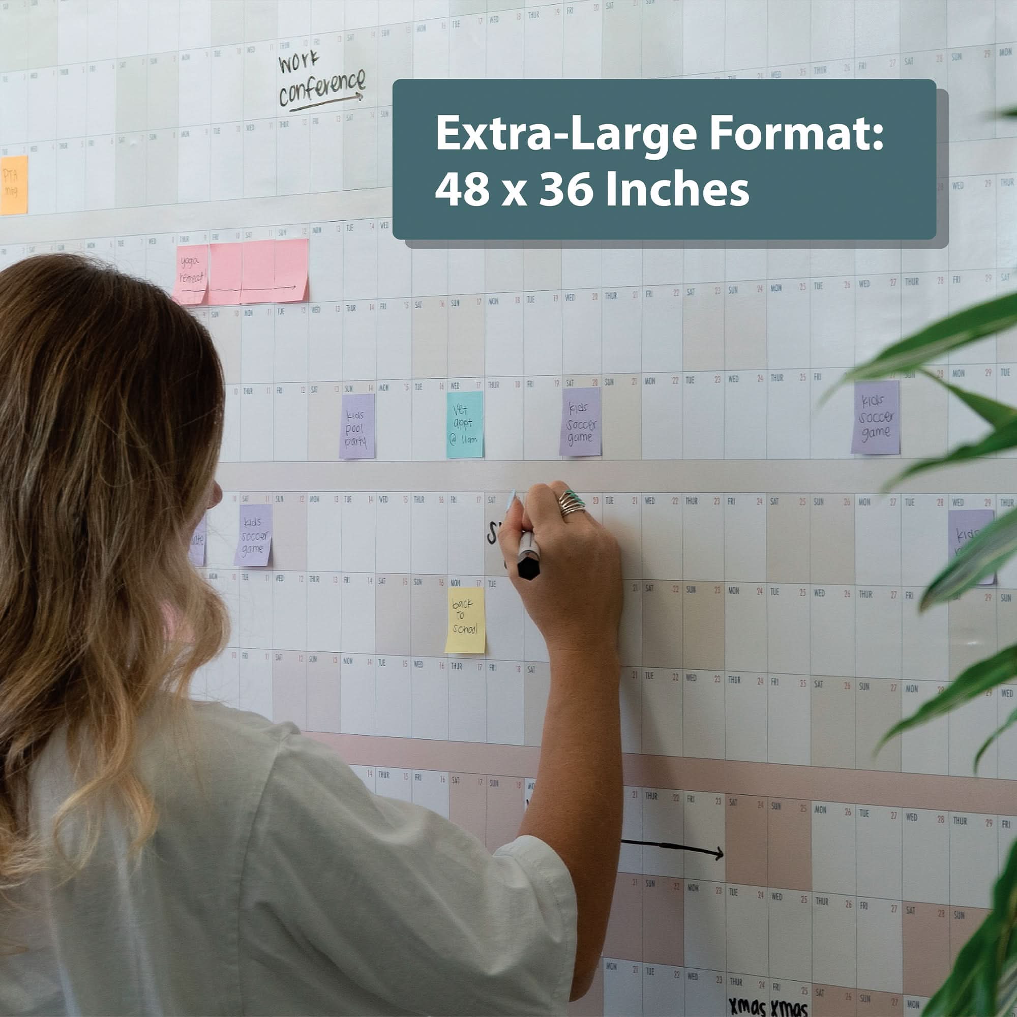 A person writes on the Eterna Goods Own The Year Calendar, a large undated wall calendar, using colored sticky notes, with a label in the top right corner that reads “Extra-Large Format: 48 x 36 Inches.”.