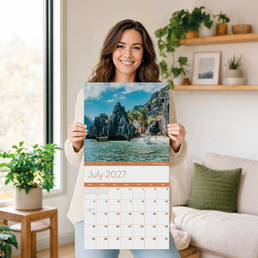A woman stands in a bright, modern living room with plants and neutral decor, holding the Eterna Goods 2027 Beaches Wall Calendar, which showcases a scenic image of limestone cliffs and turquoise water.