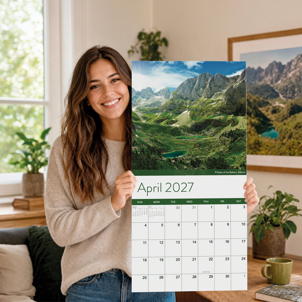 A woman stands indoors holding the 2027 Travel Inspiration Wall Calendar by Eterna Goods, which features a scenic mountain landscape photo; plants decorate the living room in the background.