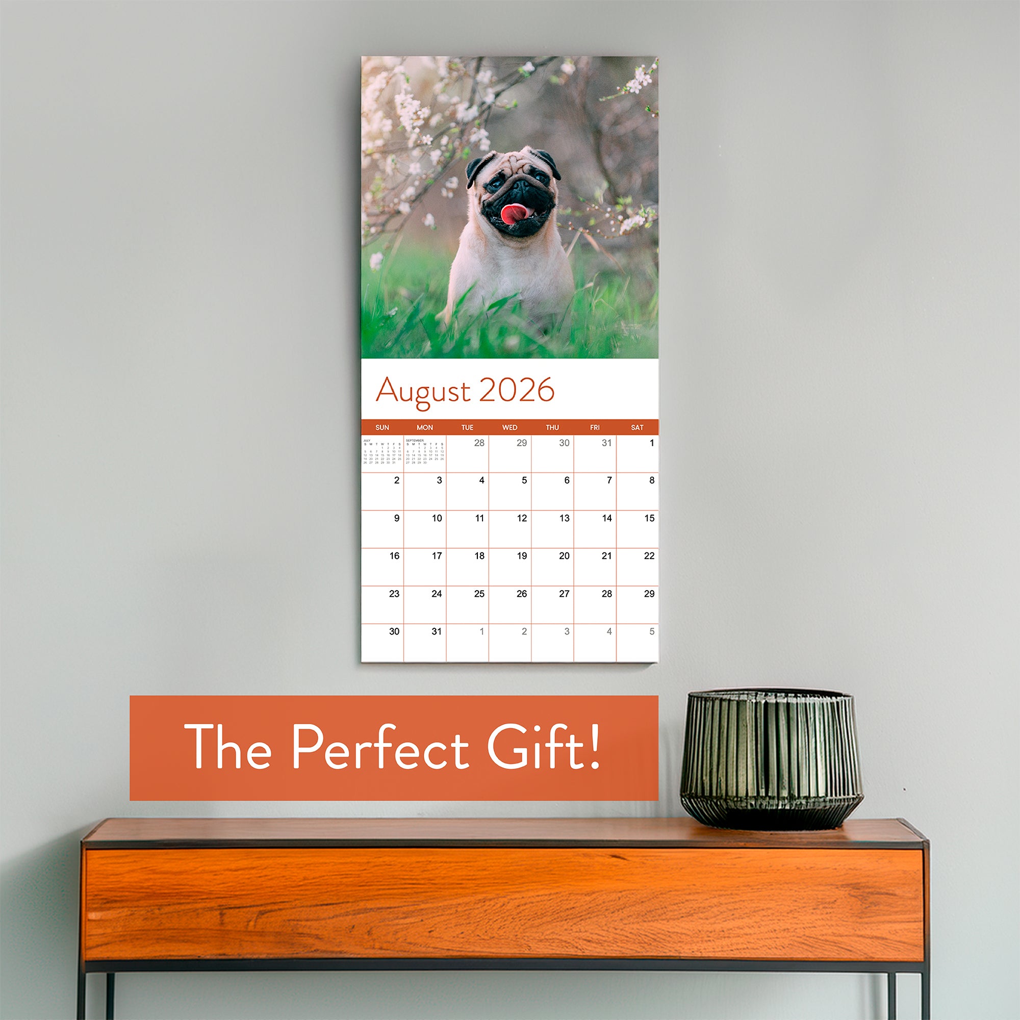 The Eterna Goods 2026 Dogs Wall Calendar, open to August with a pug photo, is displayed above a wooden table with a lamp; text reads "The Perfect Gift!"—an ideal pick for any dog calendar lover.