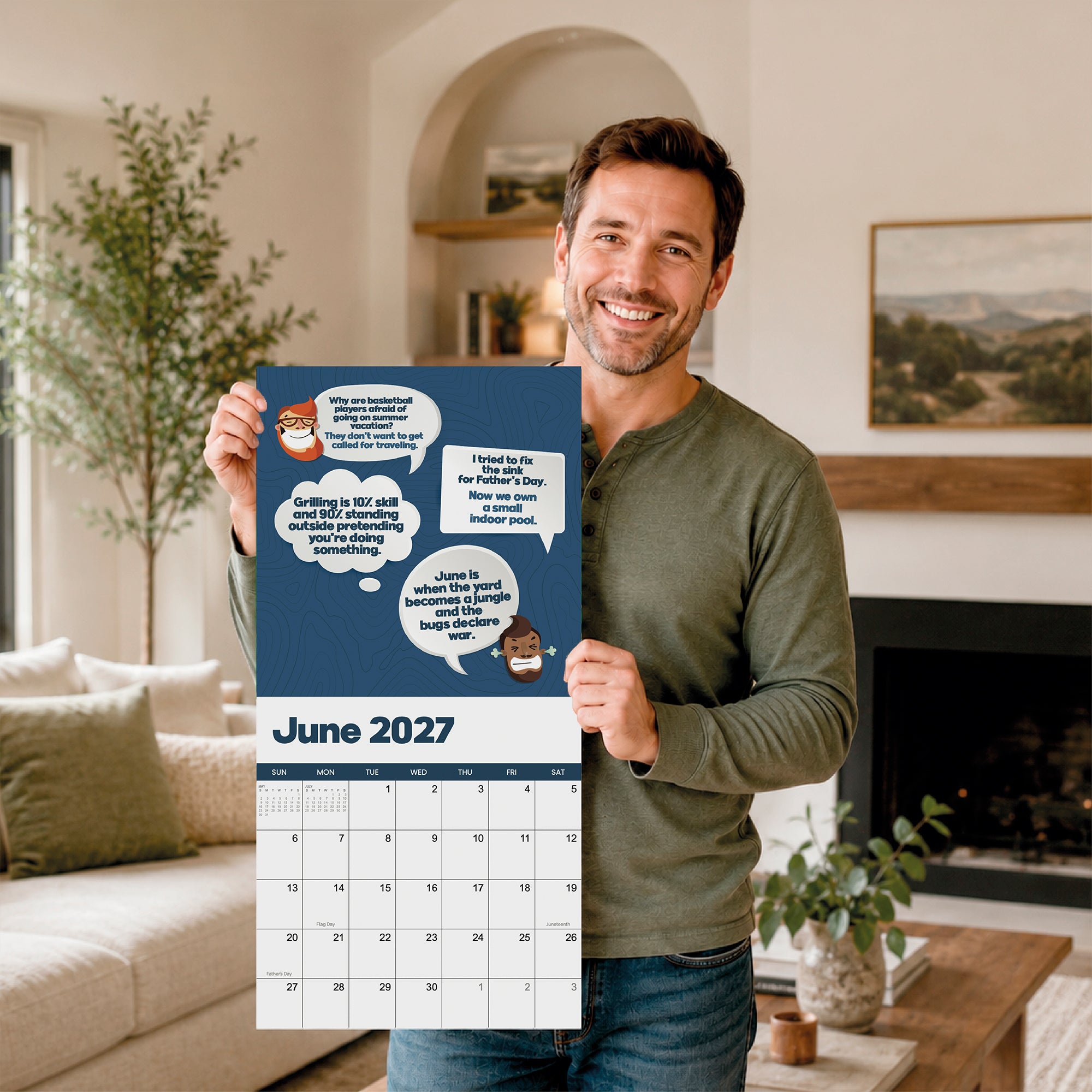 A man stands in a living room, smiling and holding a large 2027 Dad Joke Wall Calendar from That Is So Dad, featuring illustrated speech bubbles and characters at the top.