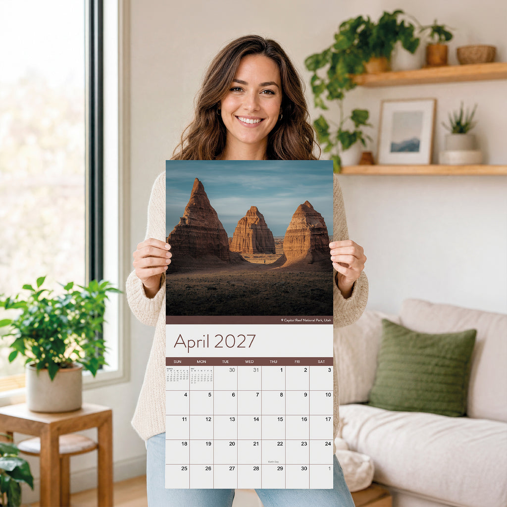 A woman stands indoors holding an Eterna Goods 2027 National Parks Wall Calendar, which displays a striking photo of rock formations. Ideal for outdoor enthusiasts and fans of National Park photography, the room around her is decorated with plants, a sofa, and shelves in the background.