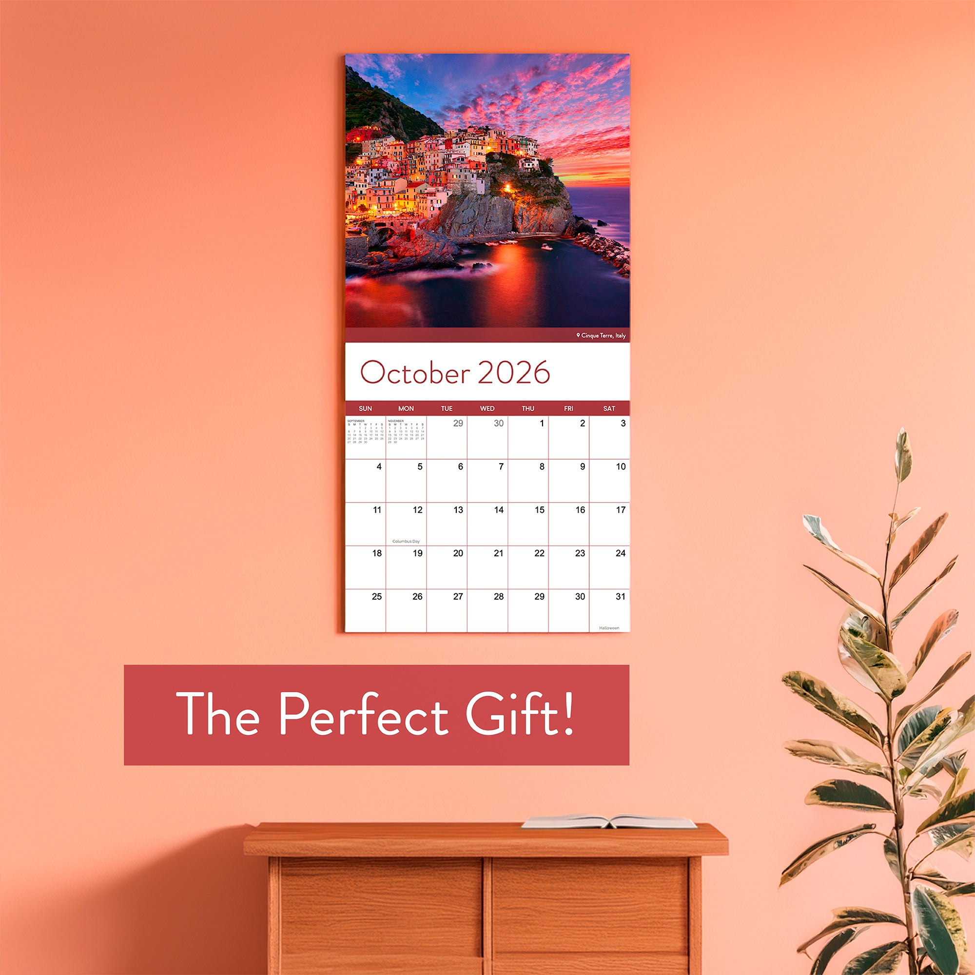 The 2026 Travel Inspiration Wall Calendar by Eterna Goods, showcasing coastal sunsets, hangs above a wooden cabinet and plant with "The Perfect Gift!" displayed on the wall.