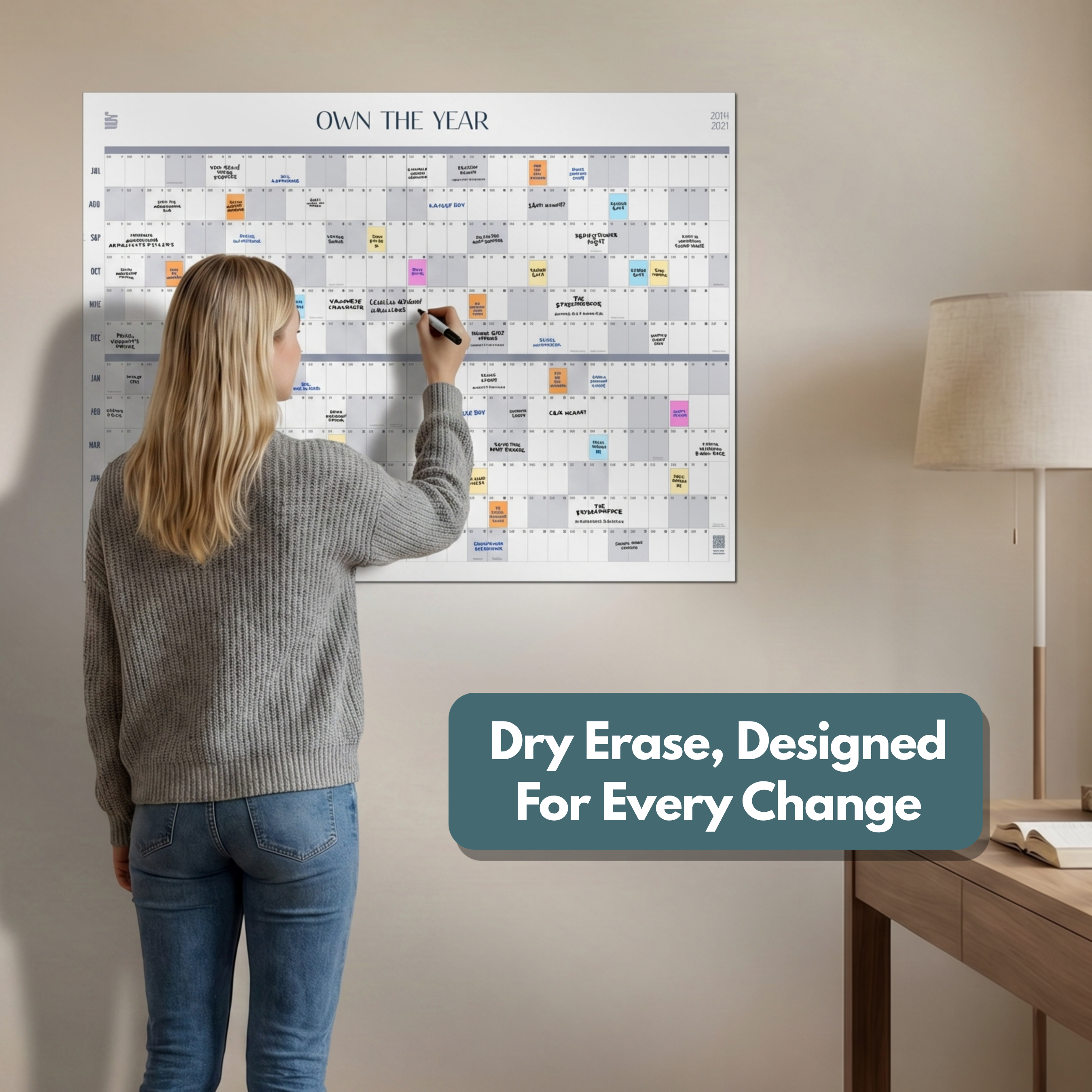 A woman writes on the Eterna Goods Midyear/Academic Own the Year 2026-2027 GIANT Dry Erase Wall Calendar (4ft x 3ft). The text reads, "Dry Erase, Designed For Every Change." Perfect for purposeful year planning.