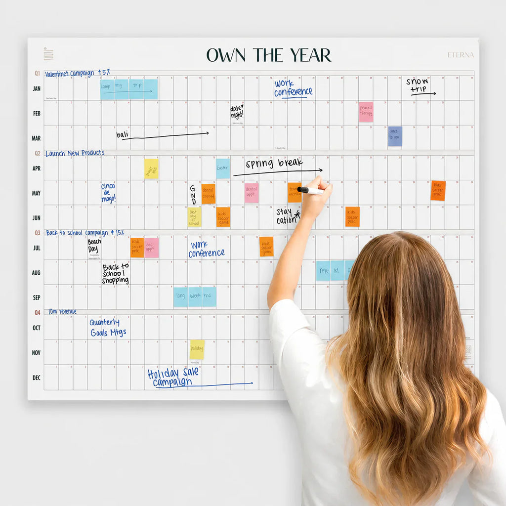 A woman writes on the Eterna Goods Own The Year Calendar, a giant dry erase wall calendar organized with color-coded sticky notes and handwritten events for each month.