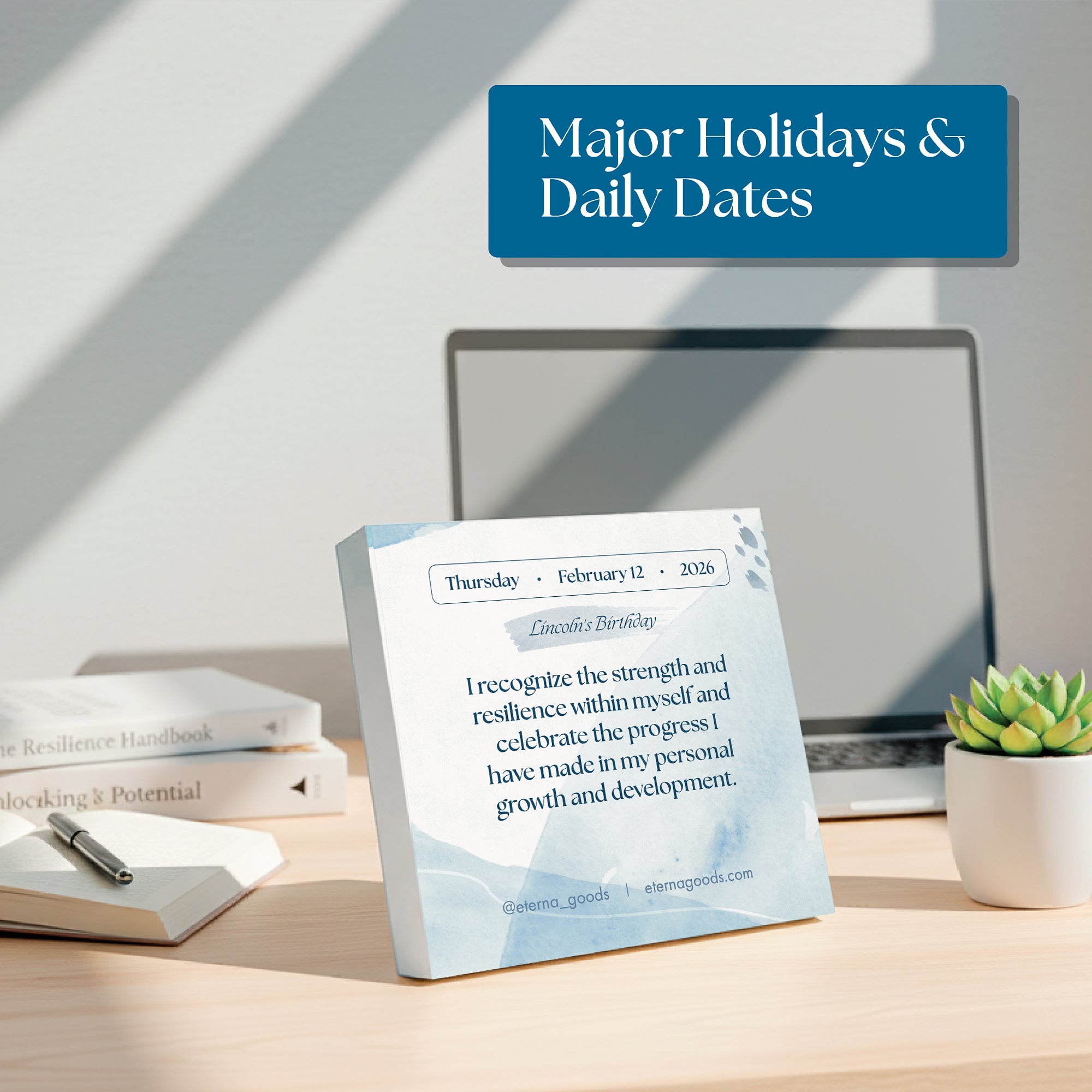 The Eterna Goods 2026 Daily Affirmations Calendar, featuring "Enoch’s Birthday" and a motivational quote, sits on a desk with a laptop, notebook, pen, and potted plant in a brightly lit office.