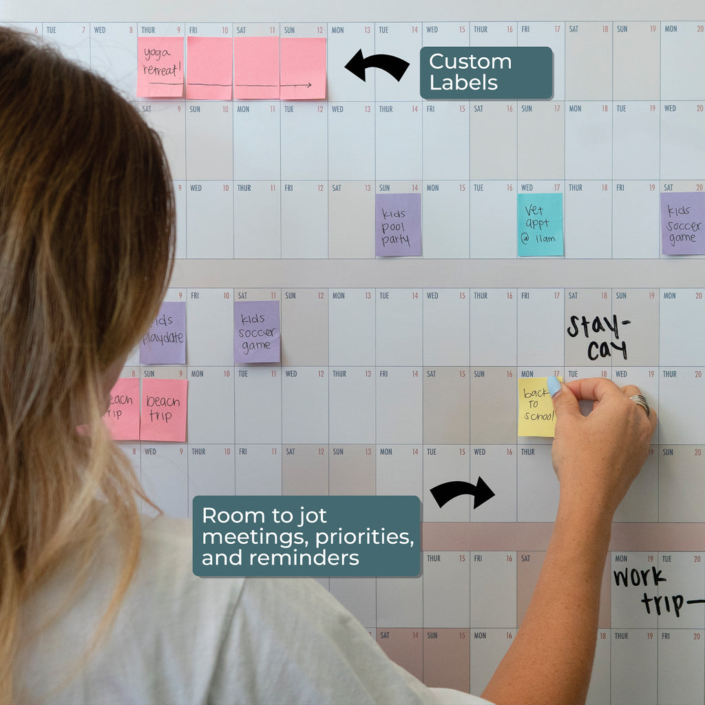 A person uses the Eterna Goods Own the Year 2026 GIANT Dry Erase Wall Calendar (4ft x 3ft) to add color-coded labels and notes for meetings, priorities, and reminders across the full year planner.