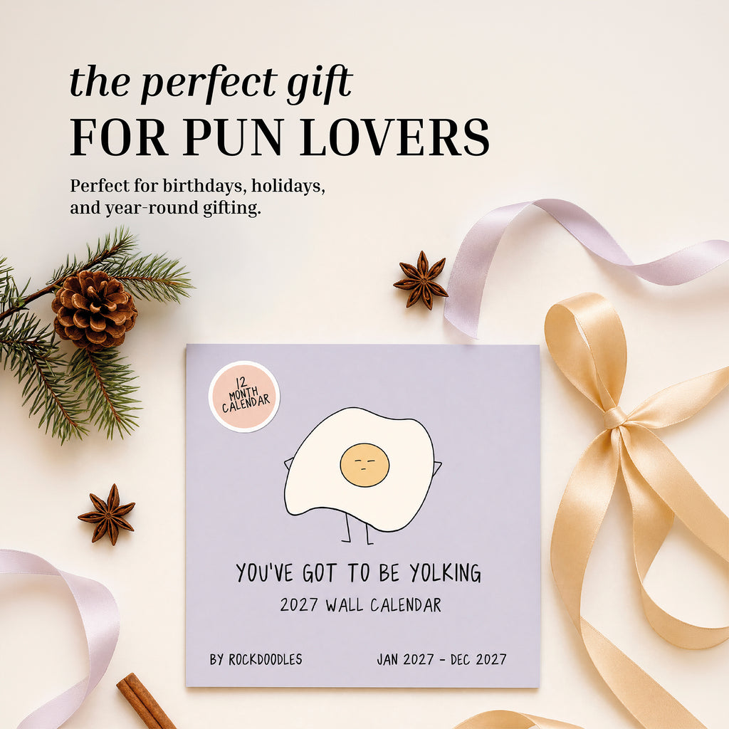 The 2027 Pun Wall Calendar by rockdoodles, featuring a cartoon egg on the cover, is displayed alongside pinecones, ribbon, and a gift bow—highlighting it as a hilarious calendar and an ideal present for fans of puns.