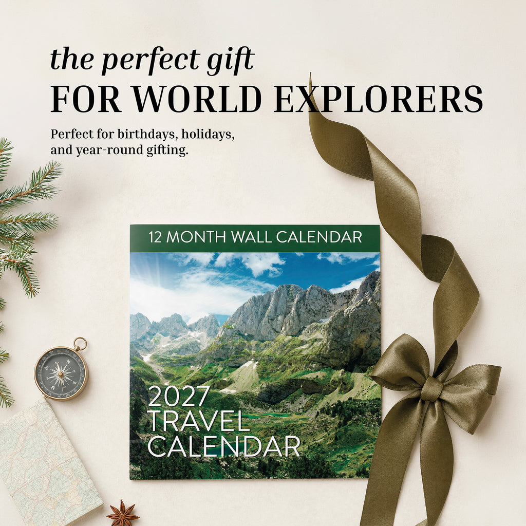 Eterna Goods 2027 Travel Inspiration Wall Calendar showcases stunning mountain photography and comes adorned with a green ribbon, compass, pen, and festive holiday greenery on a beige backdrop. The text reads: "The perfect gift for world explorers.